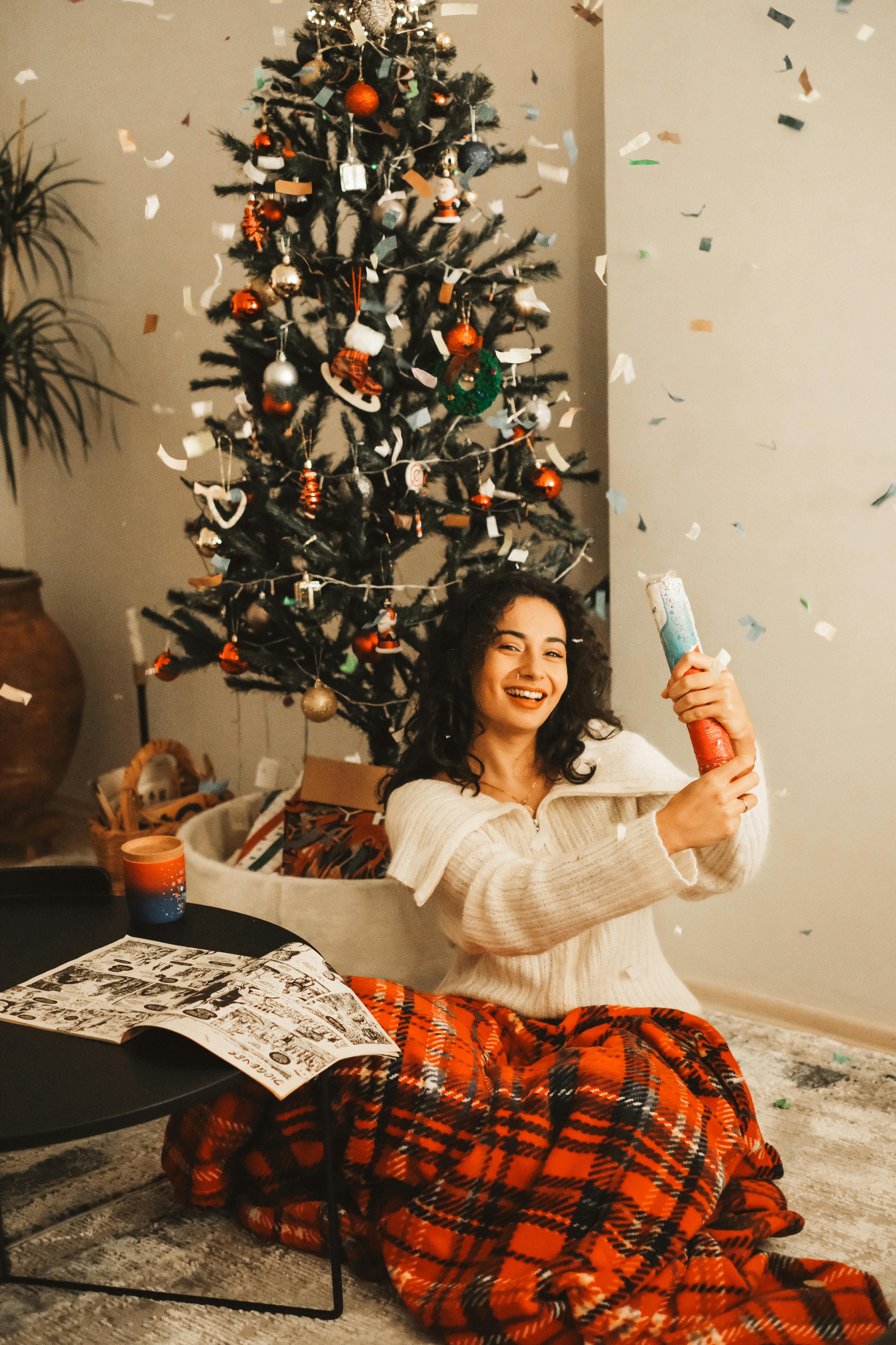 Woman celebrating the holidays indoors by a Christmas tree with confetti.