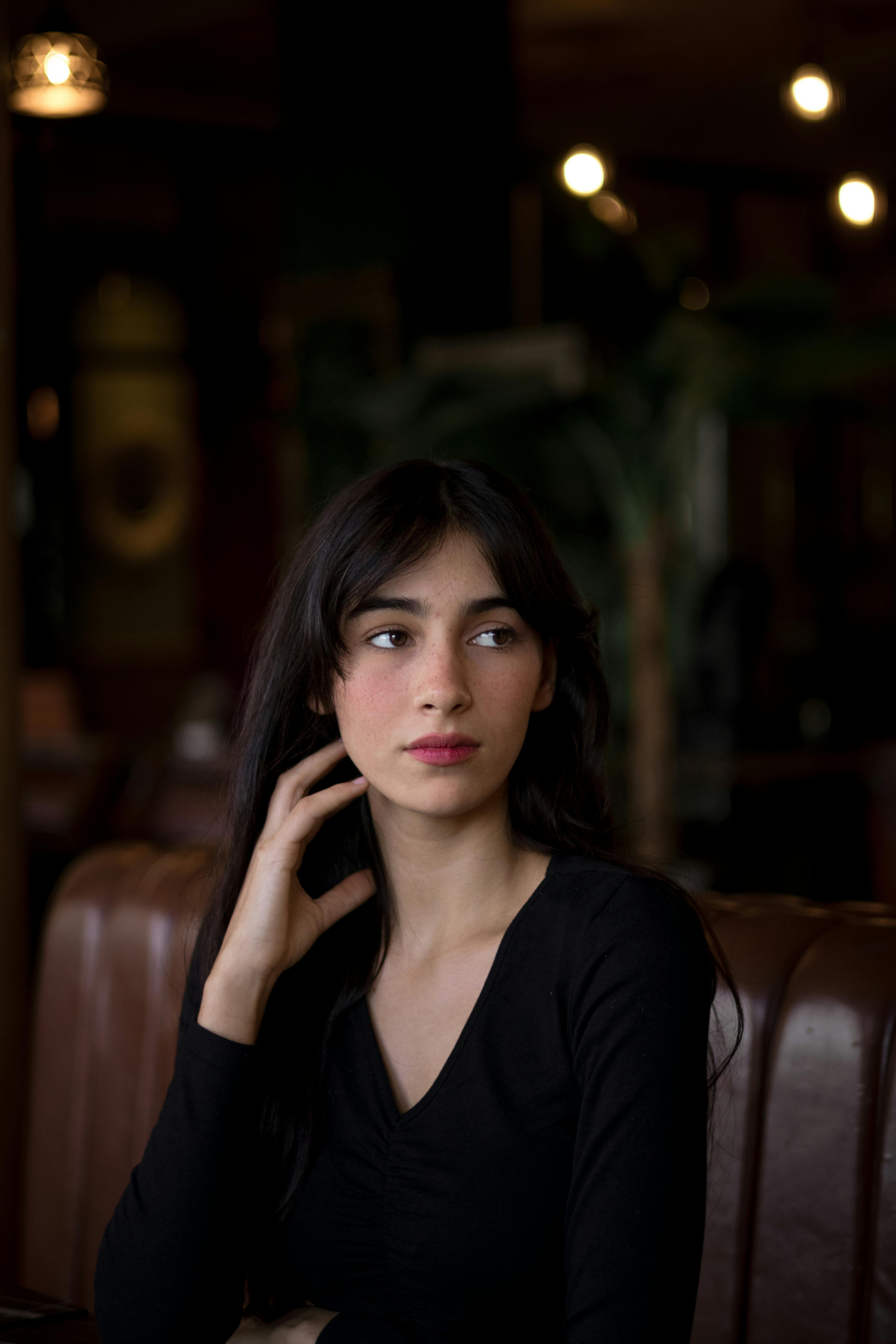 A thoughtful woman with long dark hair poses indoors wearing a black top.