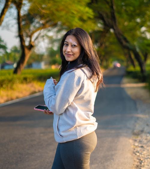Free A young woman smiles while holding a smartphone on a scenic rural road lined with trees. Stock Photo