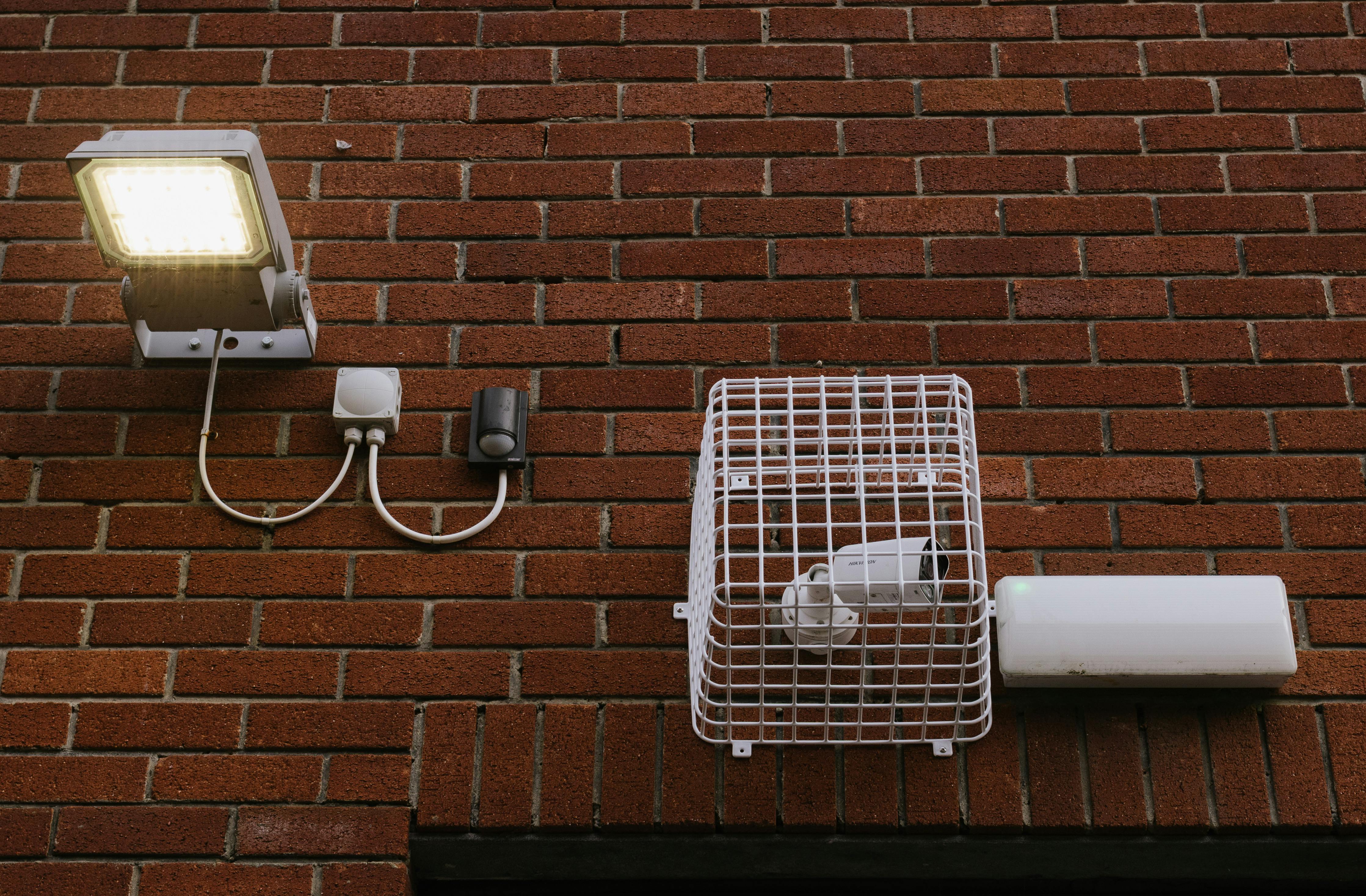 LED Lamp with a Motion Sensor and Caged Surveillance Camera on a Brick Wall