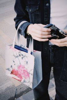 A person holds a vintage camera and a floral tote bag, walking outdoors on a sunny day.