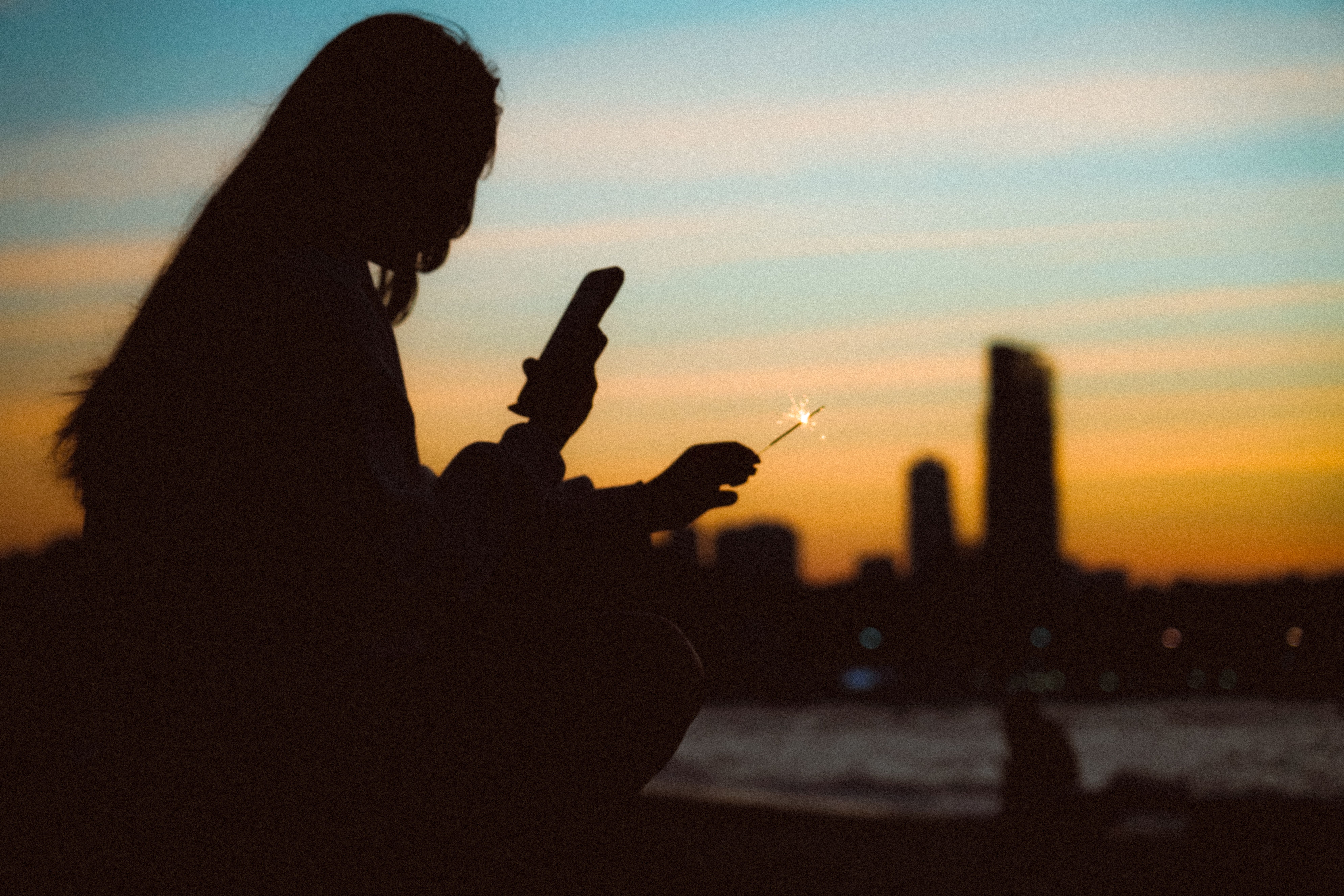 a silhouette of a man standing on a hill overlooking a city skyline at sunset, holding a phone that glows