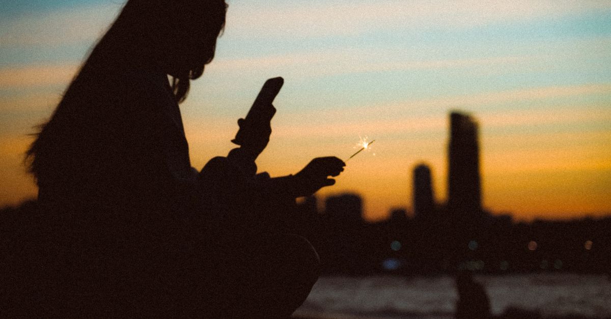 a silhouette of a man standing on a hill overlooking a city skyline at sunset, holding a phone that glows