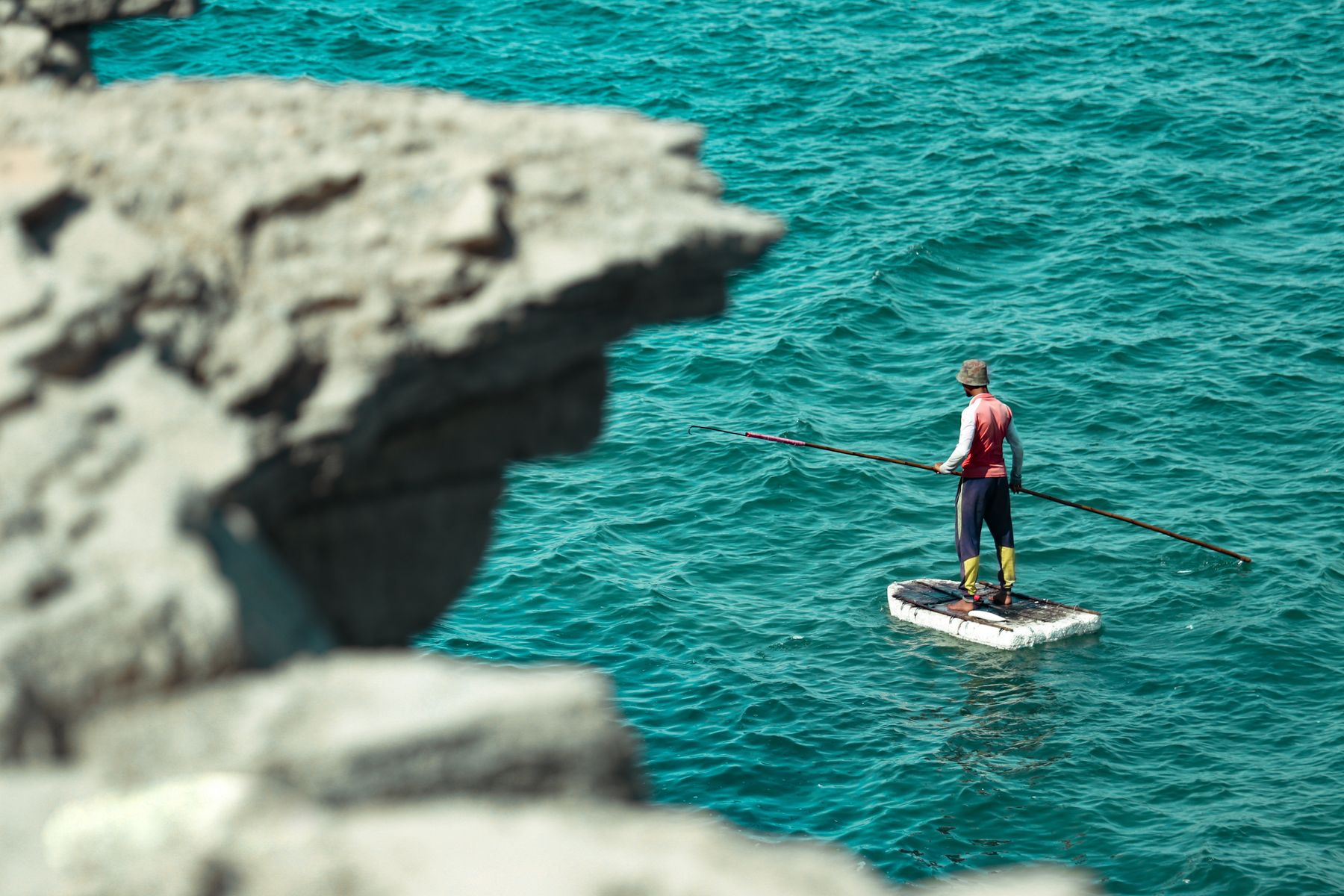 A man stands on a raft with a fishing rod in Gwadar, Balochistan, enjoying the turquoise sea and summer sun.