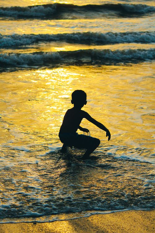 Free Silhouette of a child playing in the ocean as the sun sets, creating a golden reflection on the water. Stock Photo