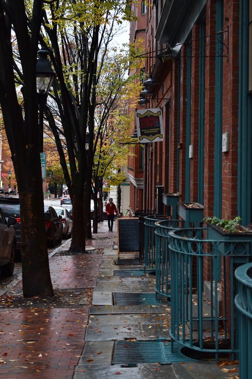 Free Charming city sidewalk scene with autumn leaves, pedestrians, and colorful buildings. Stock Photo
