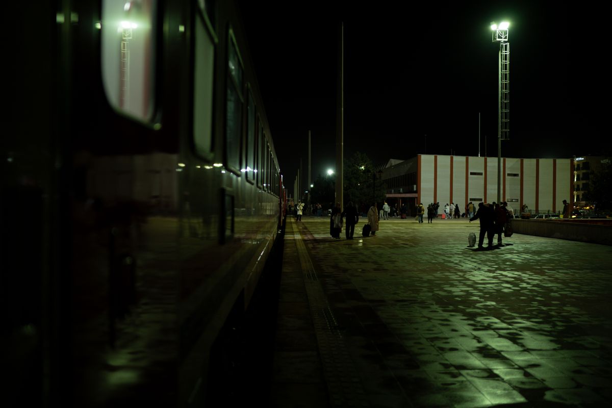 Peron de gara pe intuneric, cu tren si siluete sub lampi