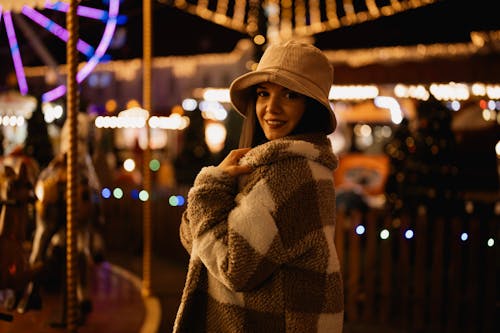 Woman Wearing a Hat and a Coat, Posing by a Carousel against City Lights at Night