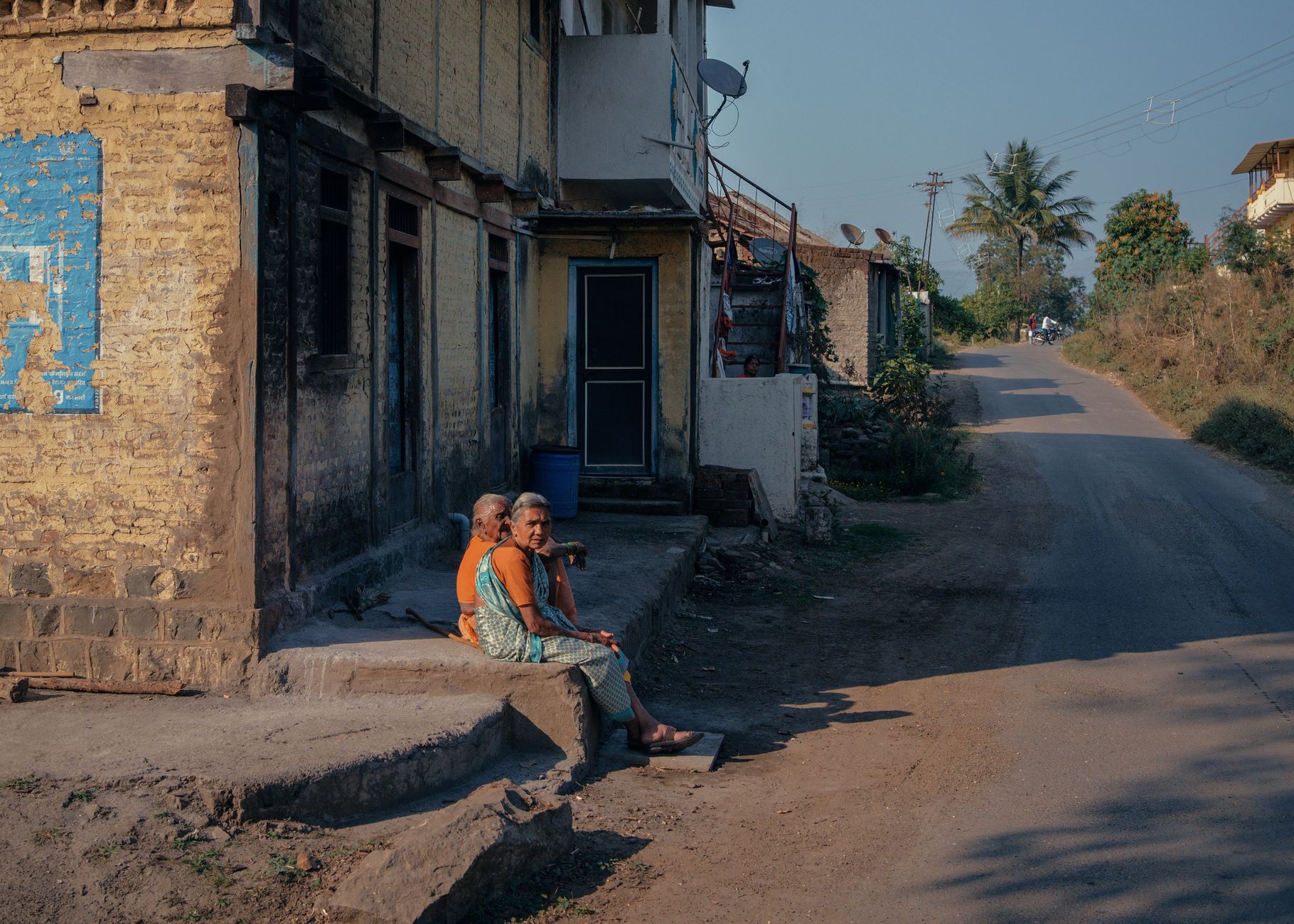 Elderly couple sitting outside a rural house in Pune's village scenery. Warm atmosphere.