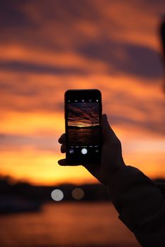 Silhouette of a hand holding a smartphone capturing a fiery sunset.