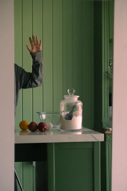 A kitchen scene with a green backdrop featuring a jar of flour, onions, and a raised hand.