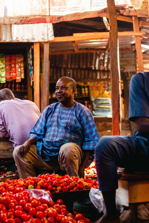 Free Cheerful vendor sitting among fresh tomatoes in a lively outdoor market setting. Stock Photo
