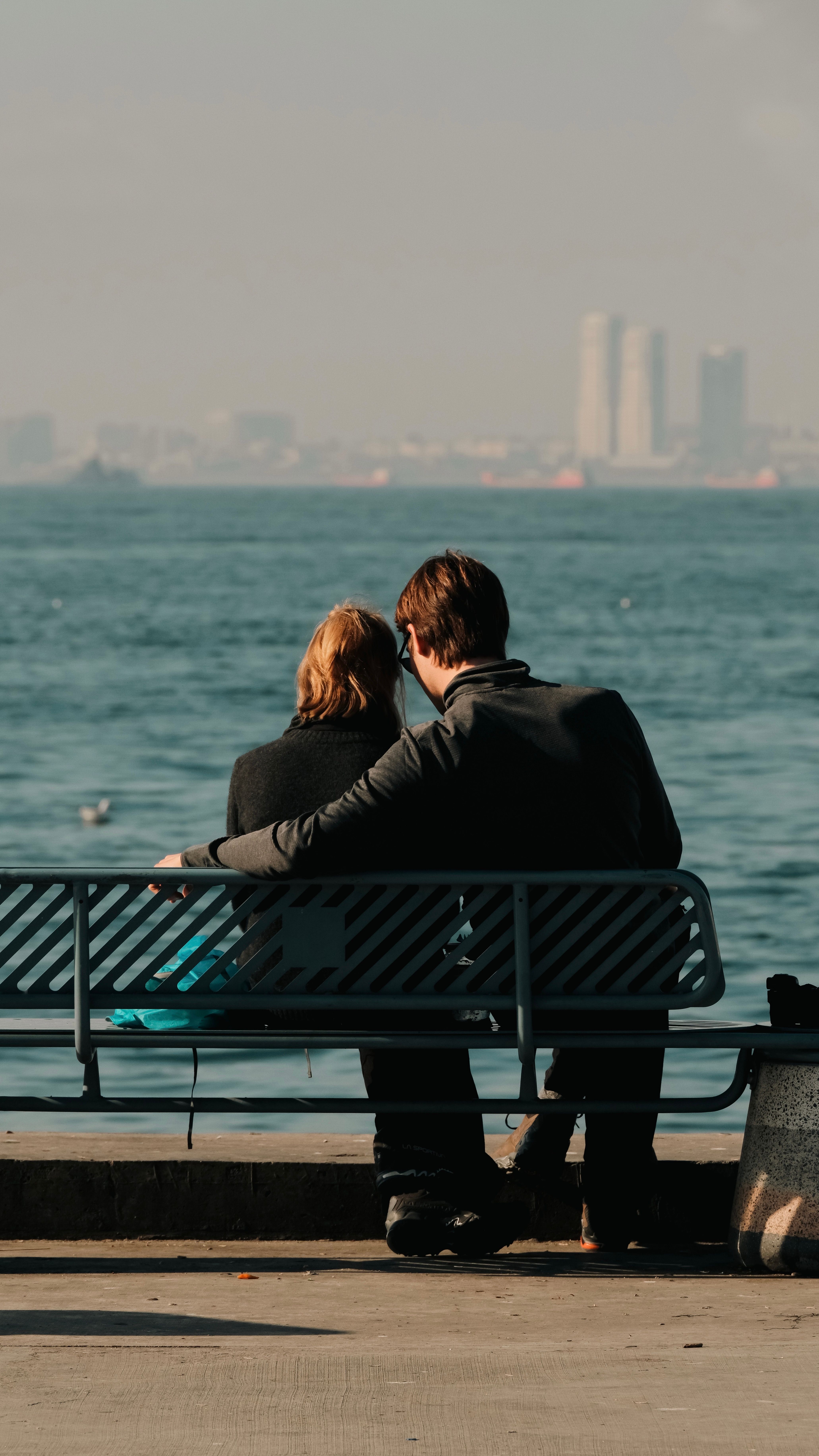 A couple enjoys a serene moment on a bench by the seaside, overlooking the cityscape.