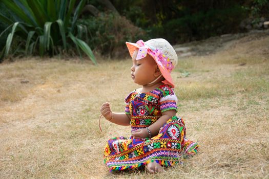 Charming baby in a colorful dress and hat sitting on grass in a serene outdoor setting.