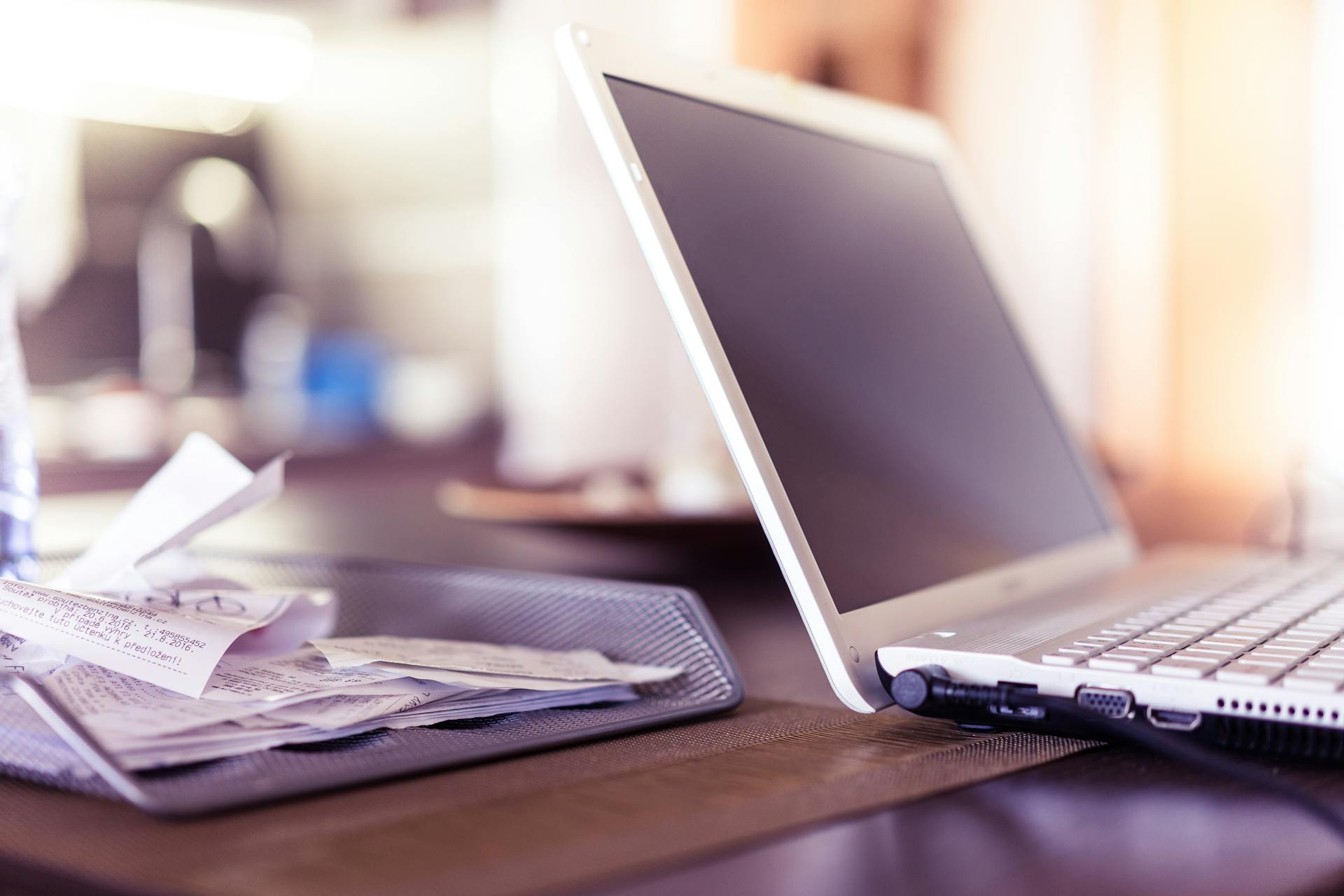 A close-up of an open laptop beside a mesh tray filled with receipts on a desk.