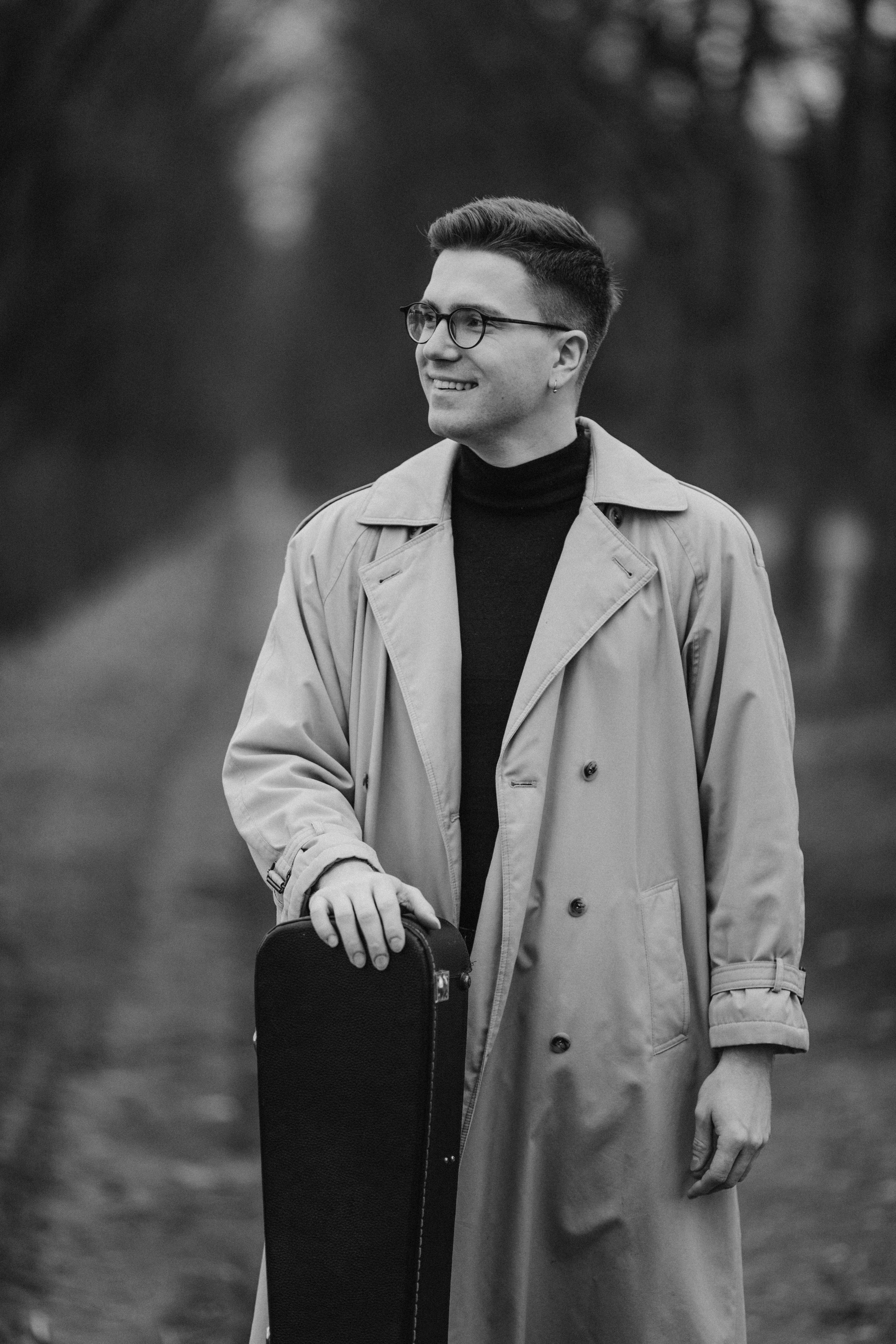 Black and white portrait of a man in a coat holding a guitar case on a wooded path.