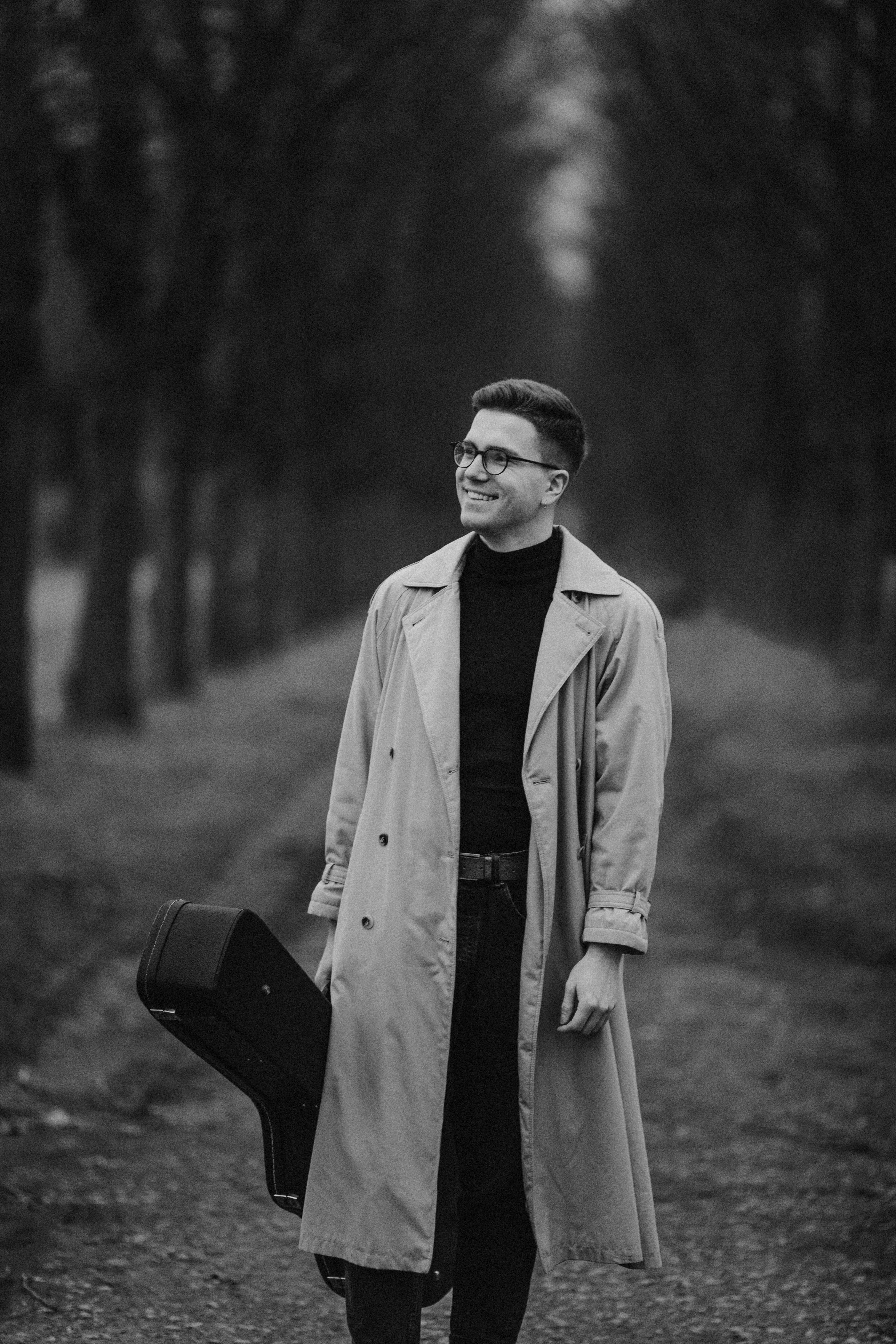 Black and white image of a man in a trench coat holding a guitar case, walking along a lined path of trees.