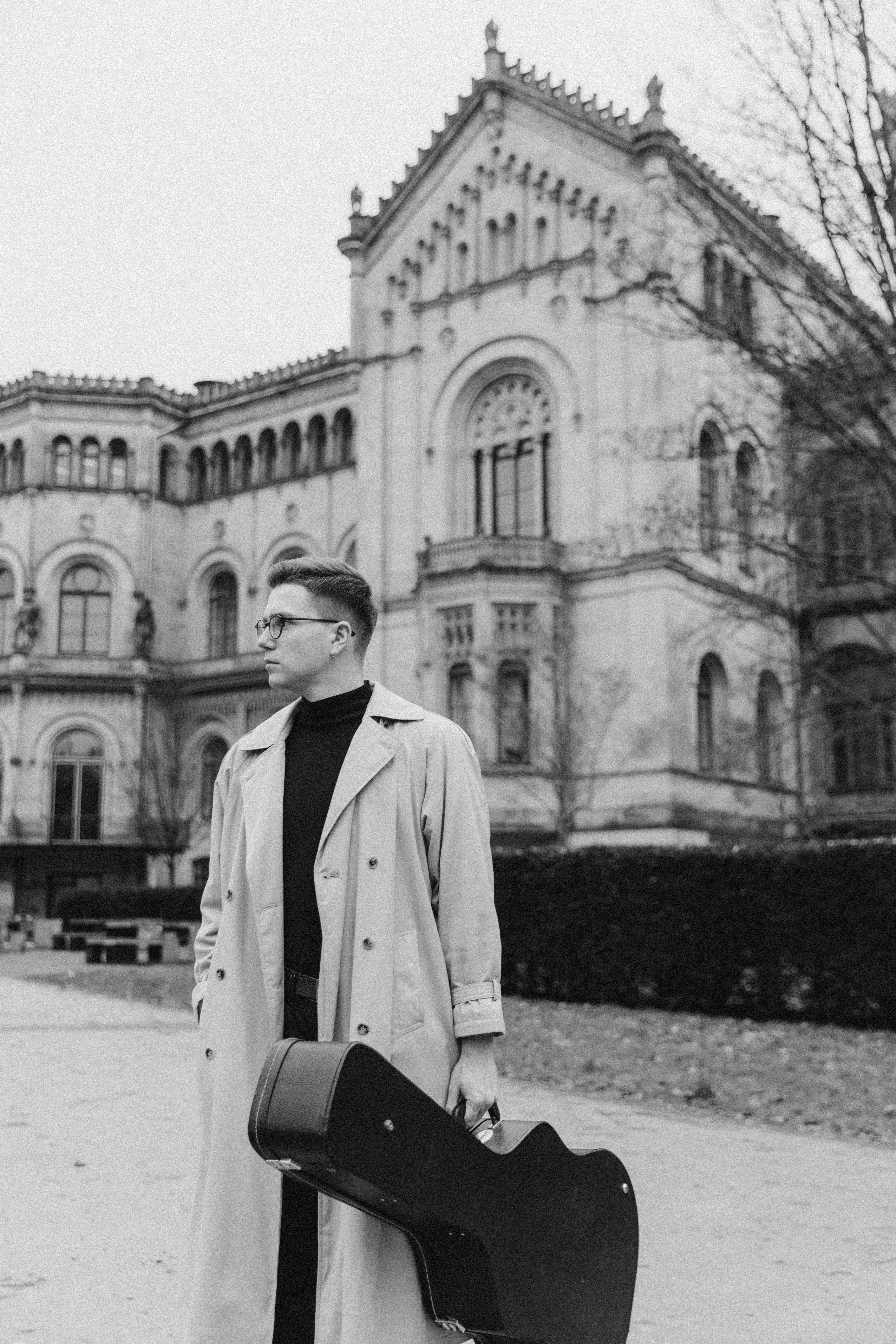 Black and white photo of a musician holding a guitar case in a cityscape.
