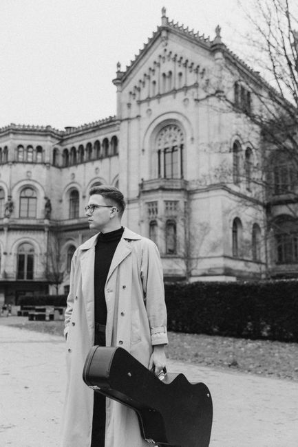 Black and white photo of a musician holding a guitar case in a cityscape.