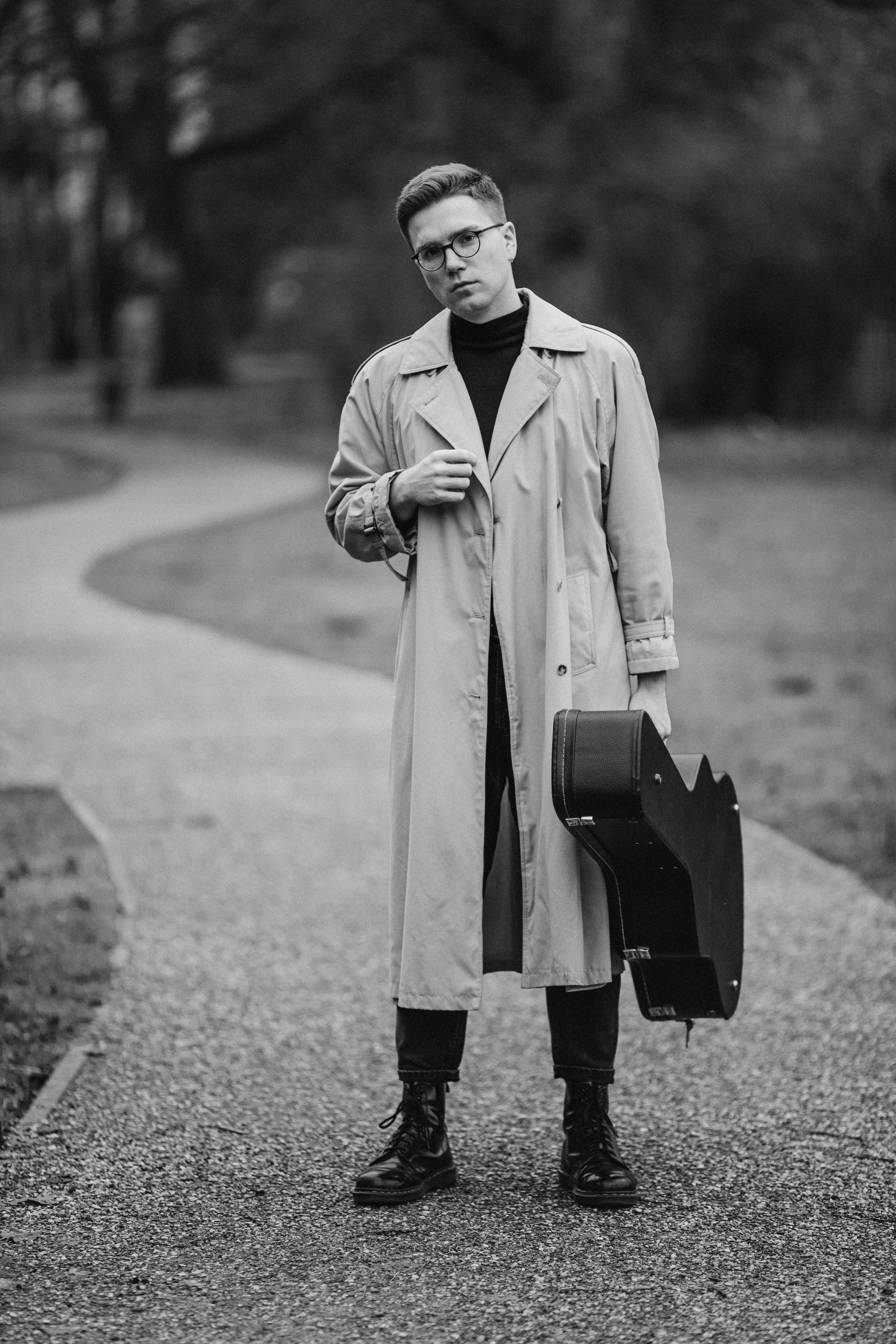 Stylish black and white portrait of a man in a trench coat holding a guitar on a park path.