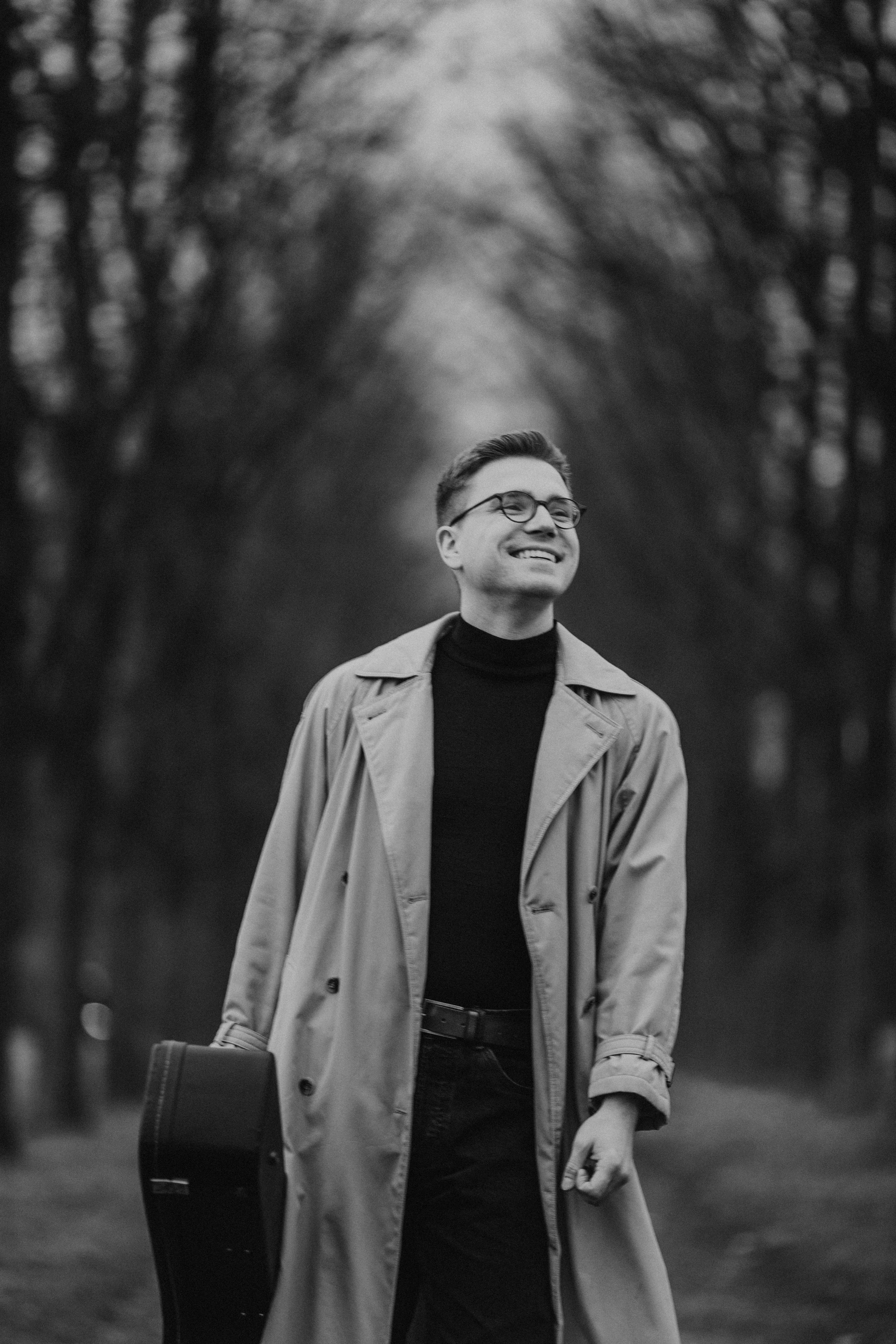 Smiling man with guitar case on a forest path, wearing a coat.