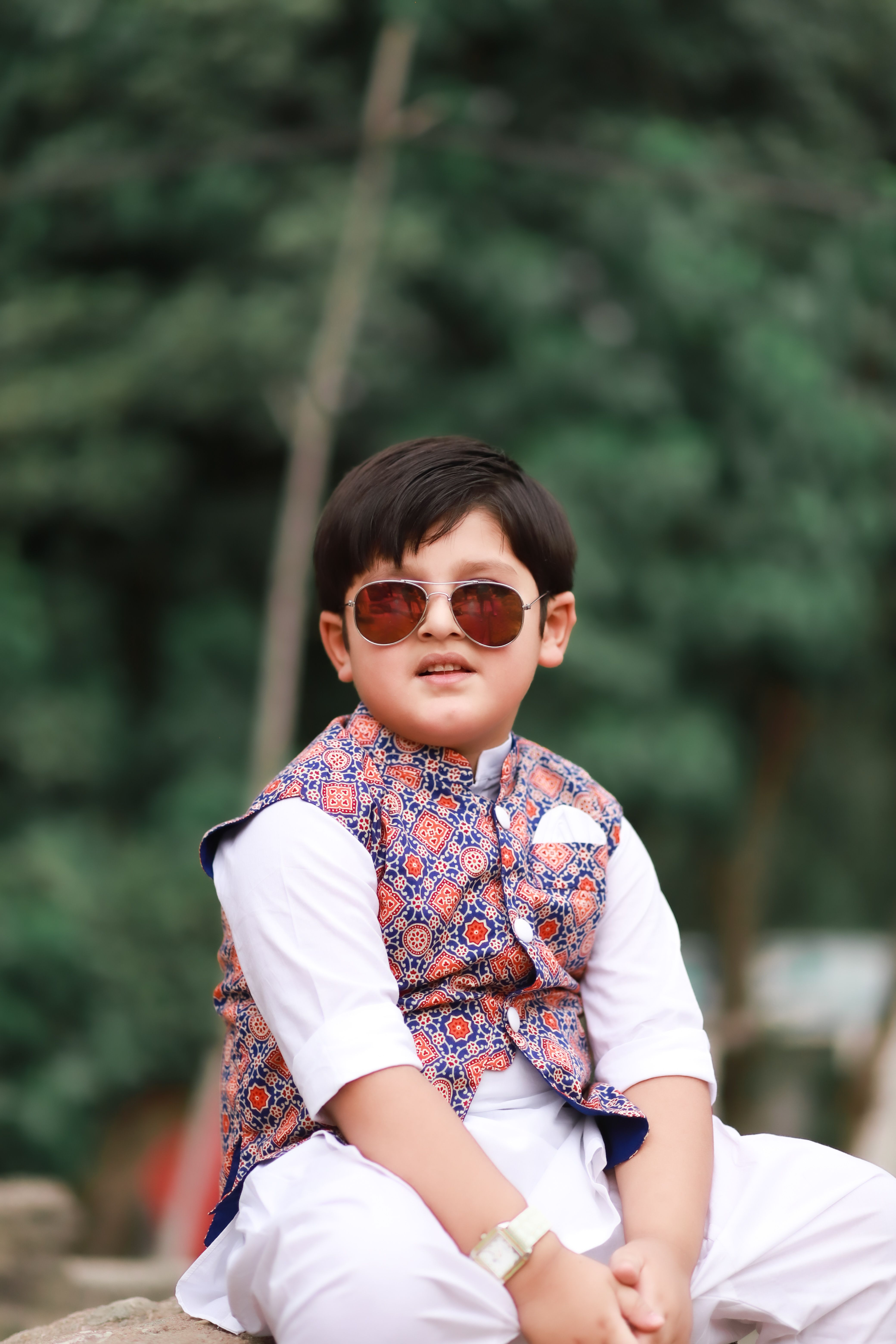 A fashionable young boy wearing sunglasses and a printed vest poses outdoors.
