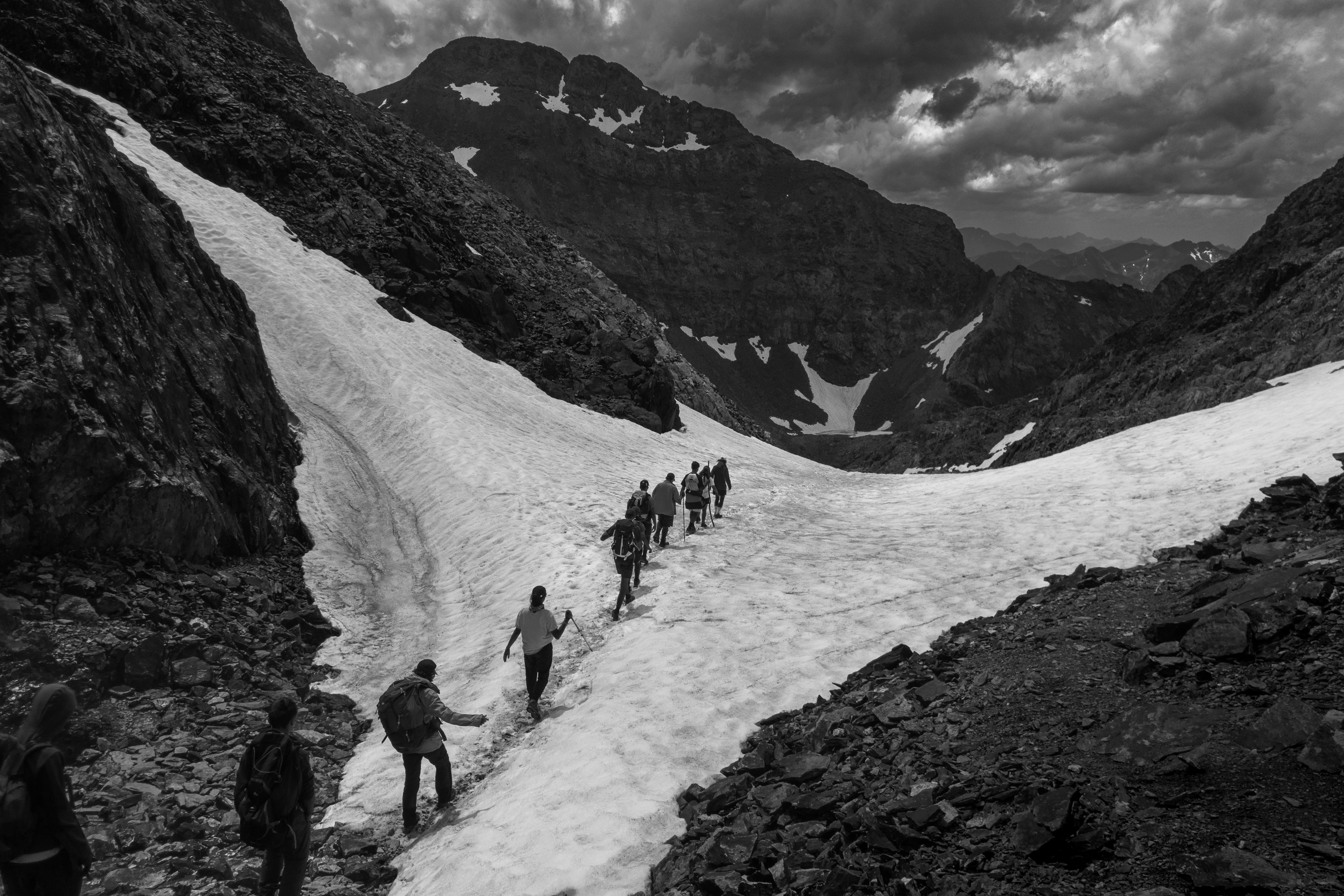 A group of people hiking across a snow-covered mountain path under dramatic clouds.