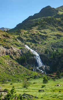 A cascading waterfall flows through a lush, green mountainside under a clear blue sky.