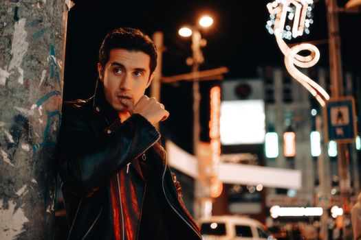 Stylish man poses in a leather jacket on bustling city street at night.