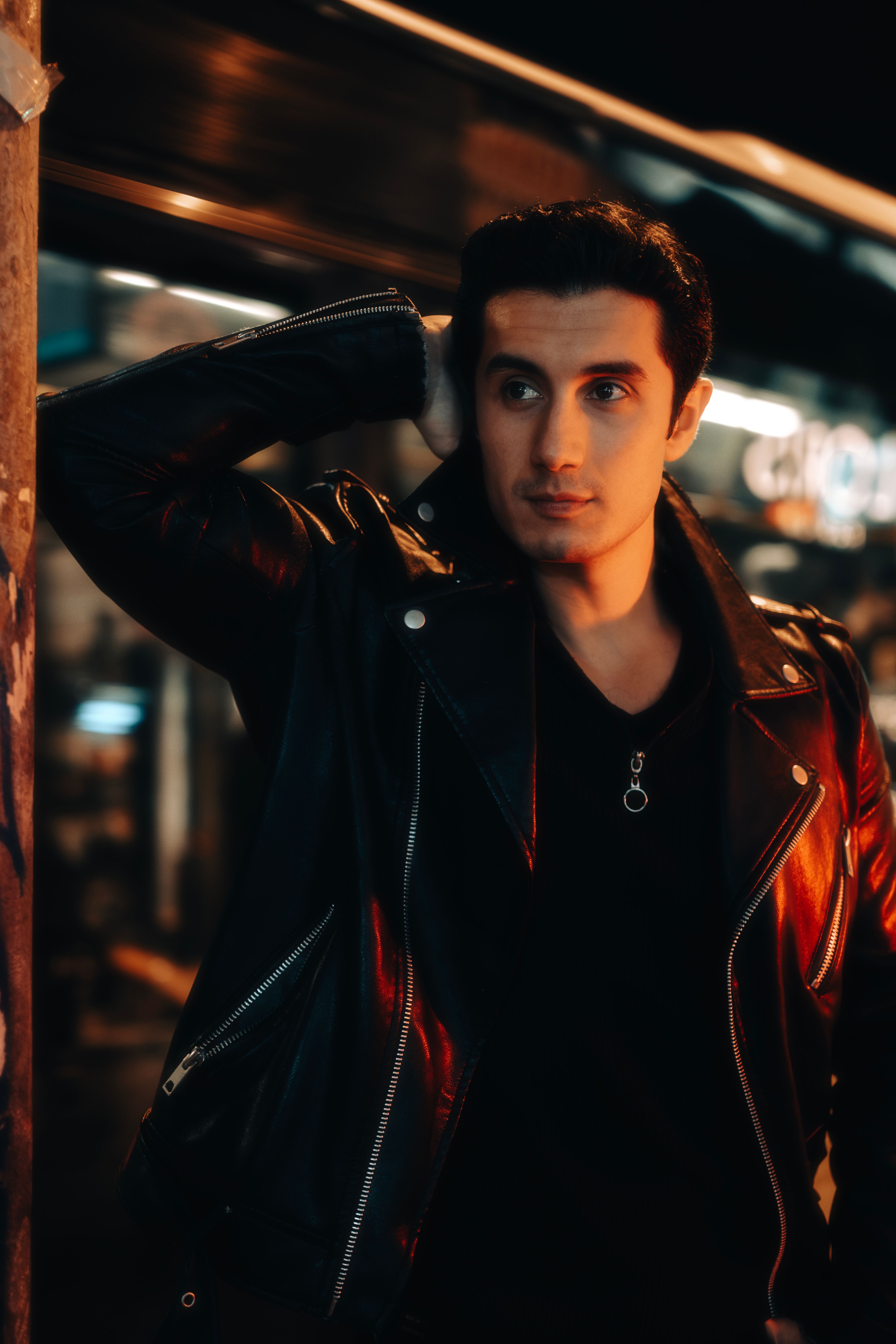 A fashionable man in a leather jacket poses confidently outdoors at night.