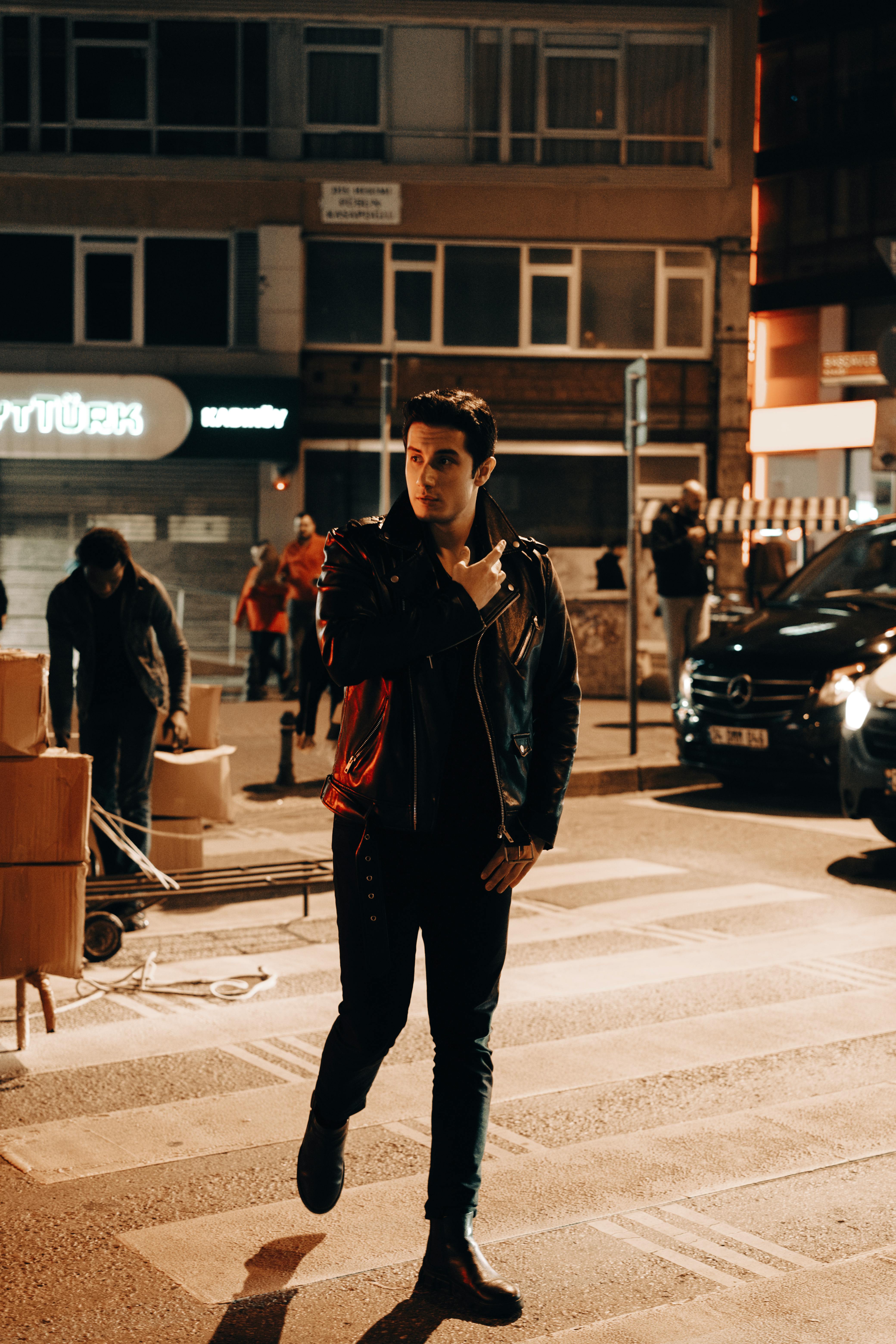 A fashionable man in a leather jacket walking across a city street at night, captured in a vibrant urban setting.
