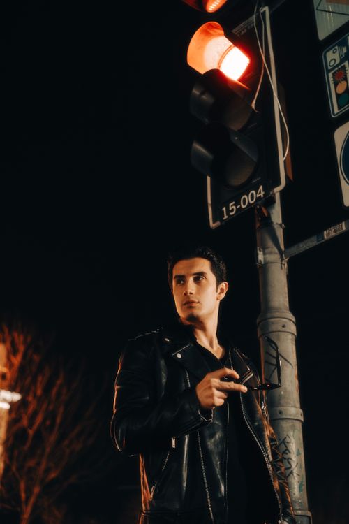Free A stylish man in a leather jacket posing by a red traffic light at night. Stock Photo
