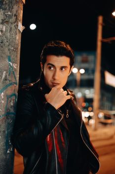 Striking nighttime portrait of a man in a leather jacket leaning against a graffiti-covered pole.