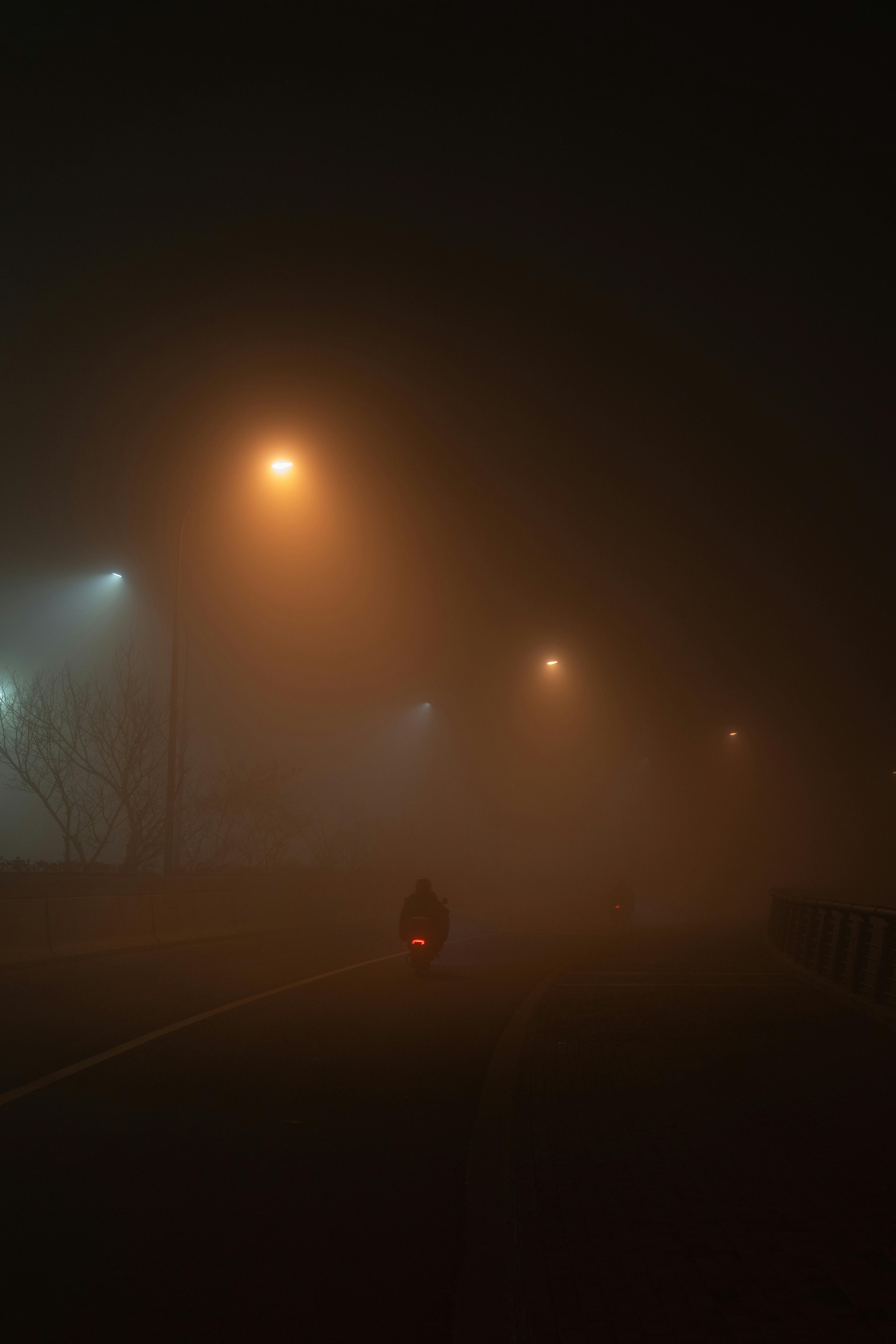 A silhouette on a motorcycle rides through foggy streets illuminated by street lights at night.