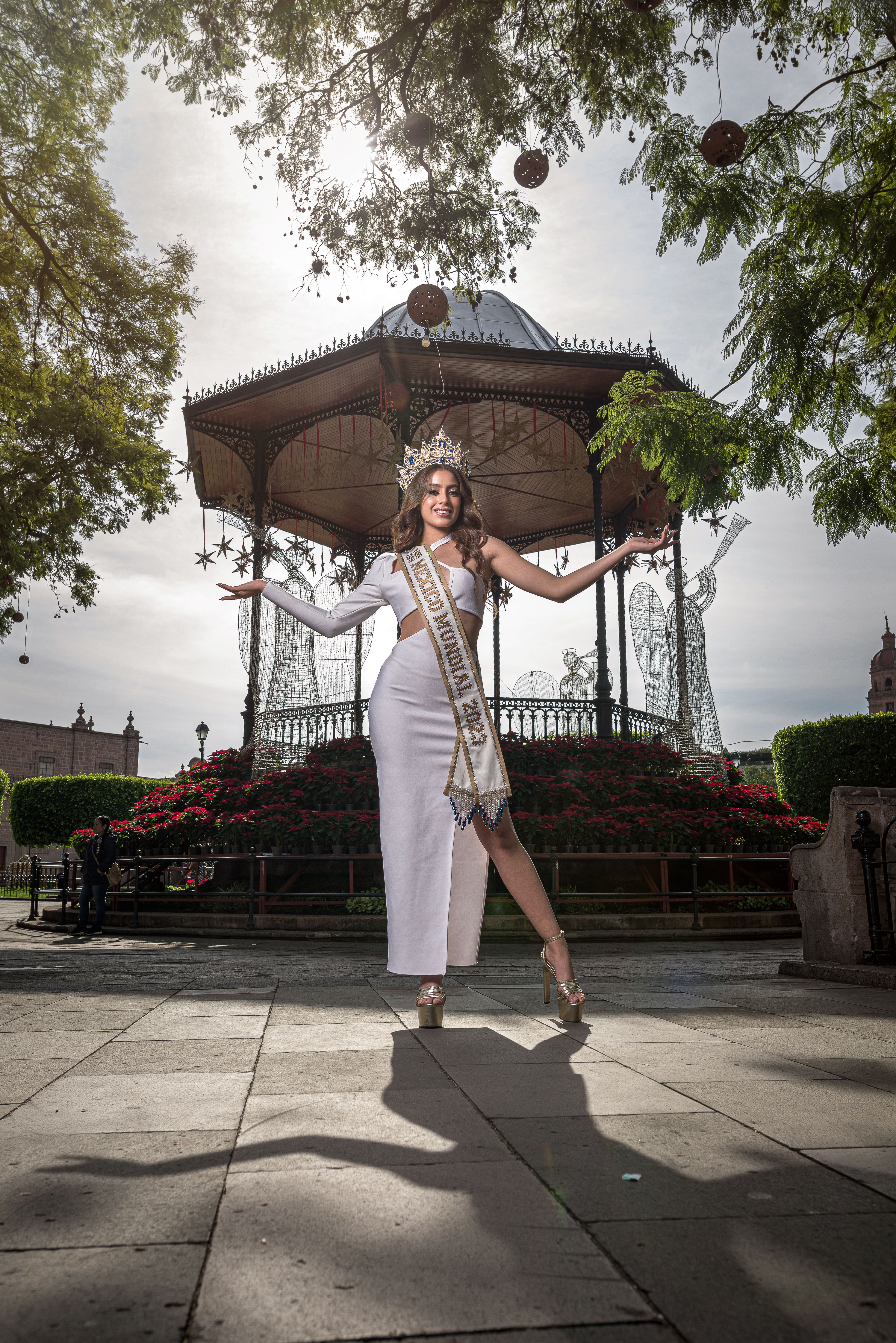 A young beauty queen in a white dress and crown poses gracefully in a city park setting.