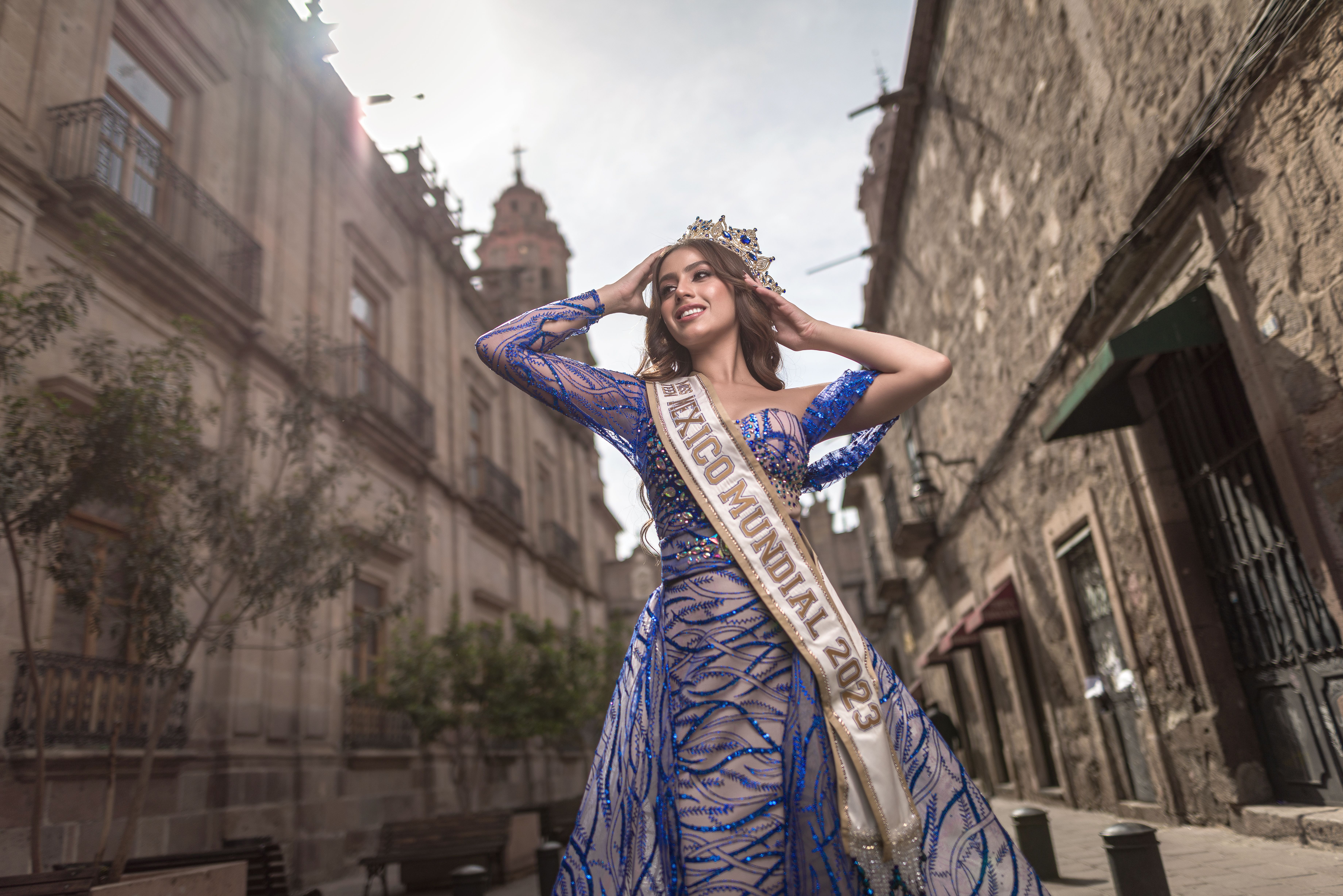 A young woman in a glamorous blue dress with a sash walks confidently through a historic urban area.