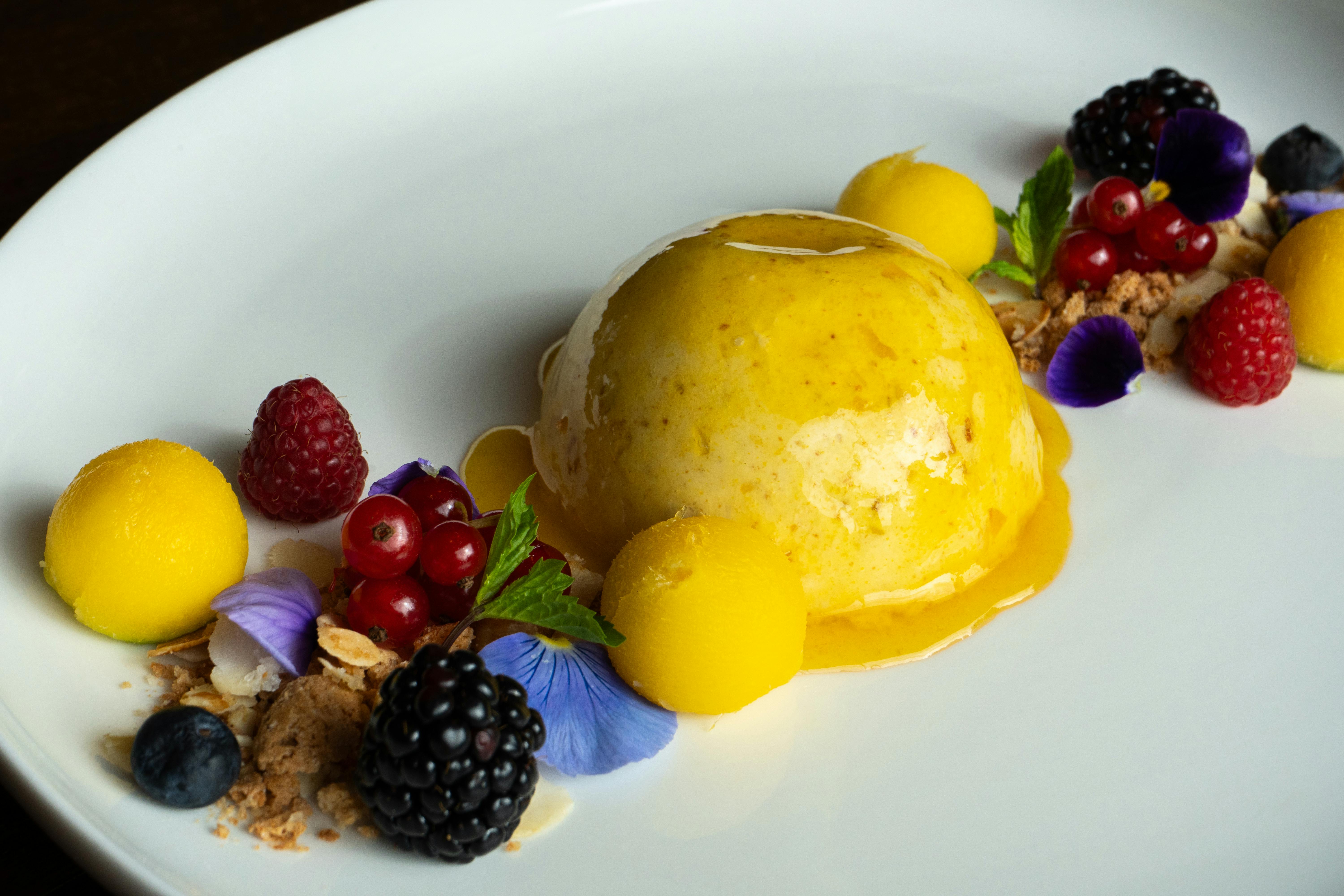 A delicious mango mousse surrounded by fresh berries and edible flowers on a white plate. | BocaVibe