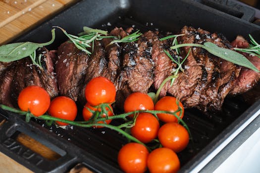 Sliced grilled steak accompanied by cherry tomatoes and fresh rosemary in a cast iron pan.