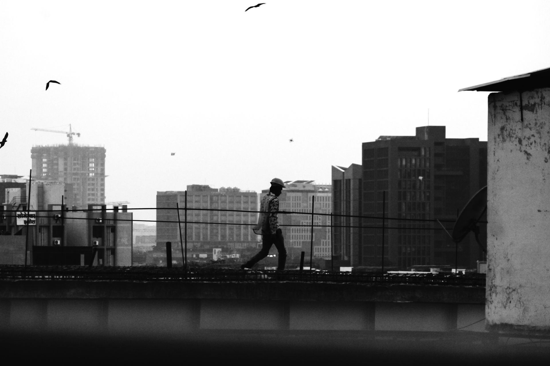 Black and white photo of a construction worker walking on a rooftop silhouette in an urban cityscape.