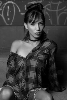 Black and white portrait of a woman in a flannel shirt, sitting against a graffiti wall.