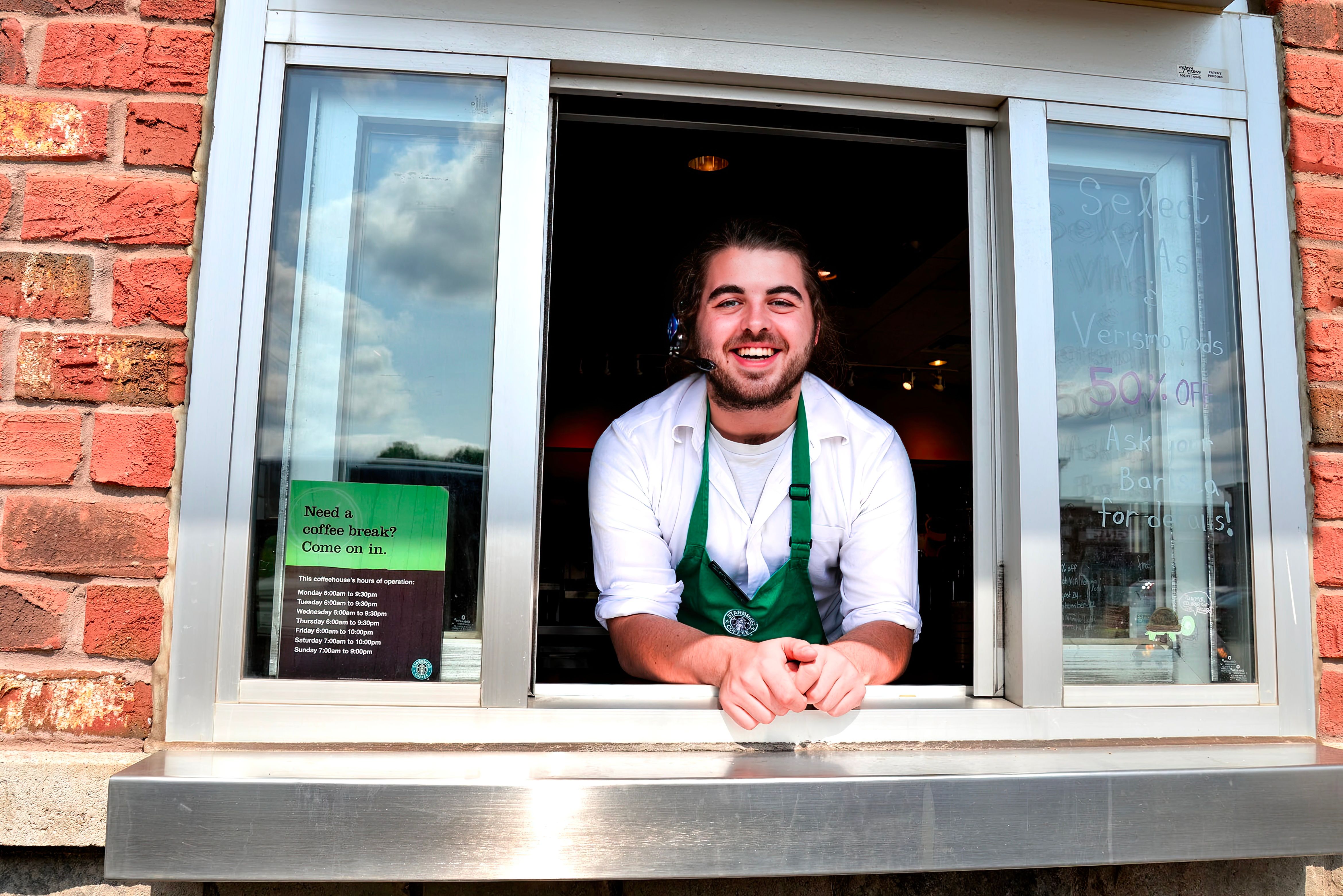 Smiling barista greeting customers at a café drive-thru window with a cheerful demeanor.