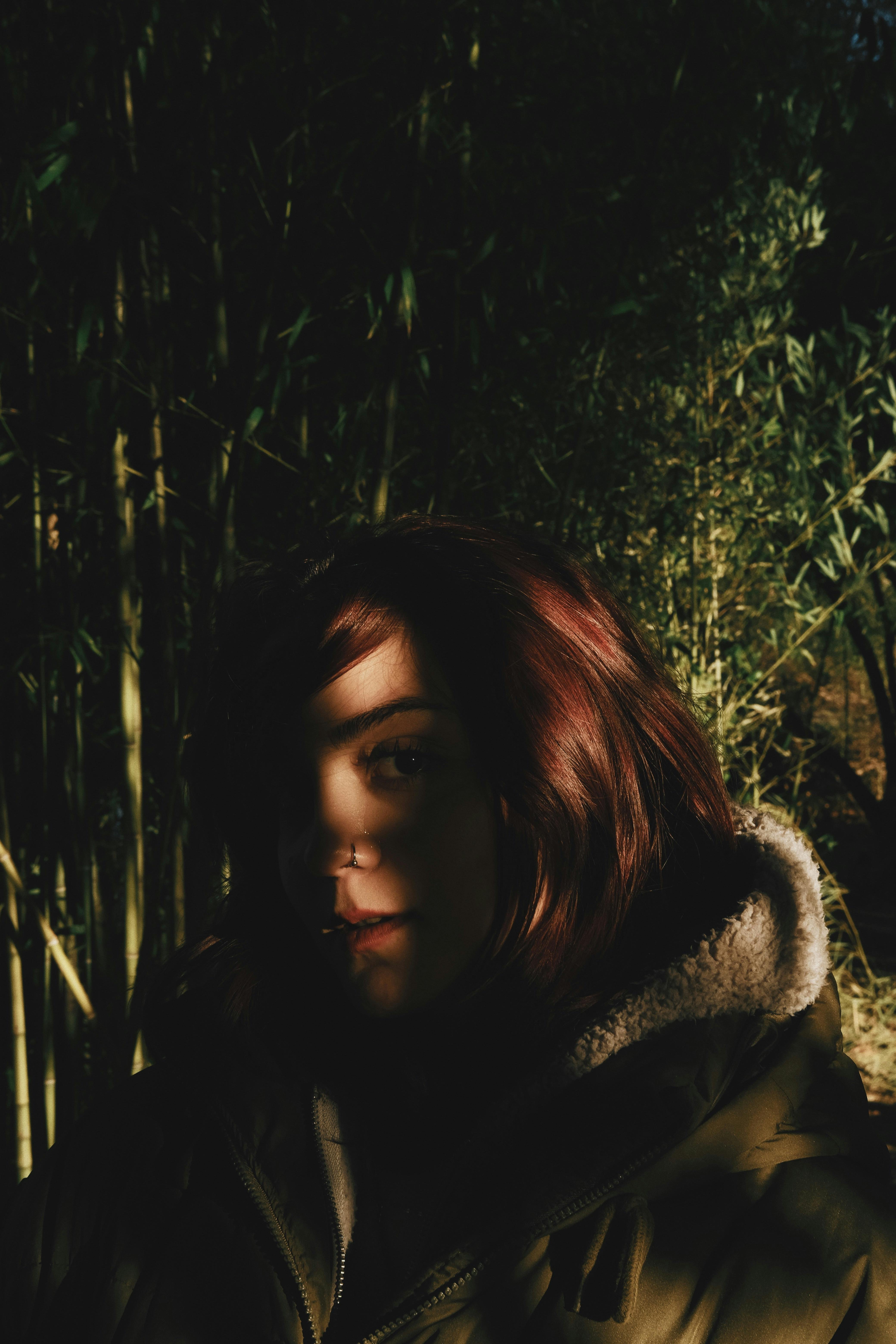 A shadowed portrait of a woman with red hair in a forest, highlighting a moody and introspective atmosphere.
