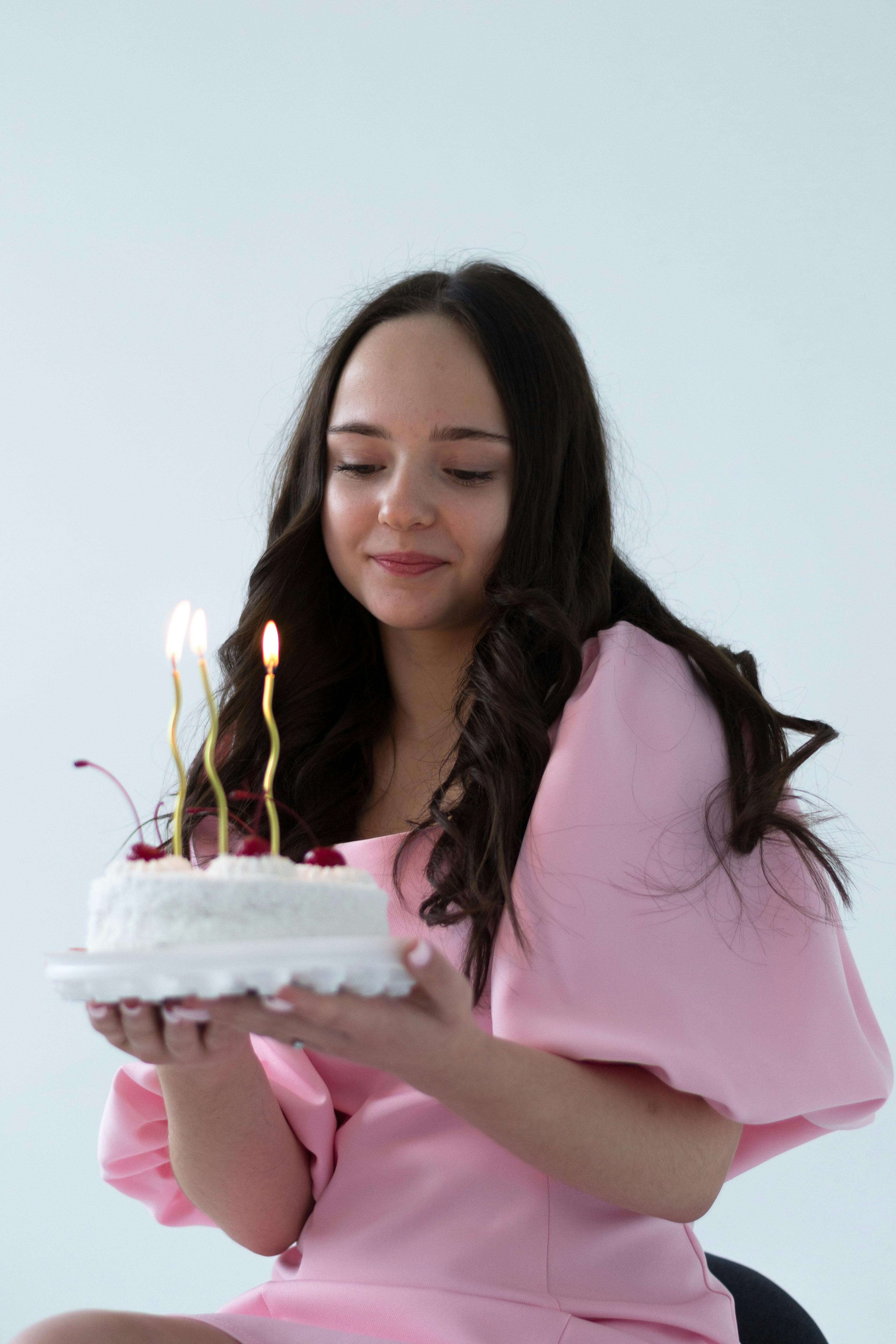 Smiling woman holding a birthday cake with candles, wearing a pink dress and celebrating indoors.