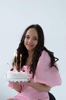 Young woman in elegant pink dress holding a birthday cake with lit candles, smiling.