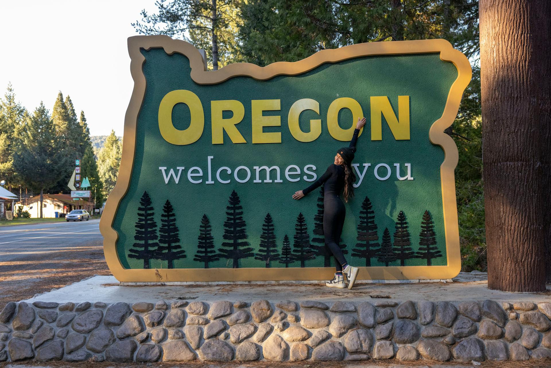 A woman joyfully poses with the iconic Oregon welcome sign on a sunny day.