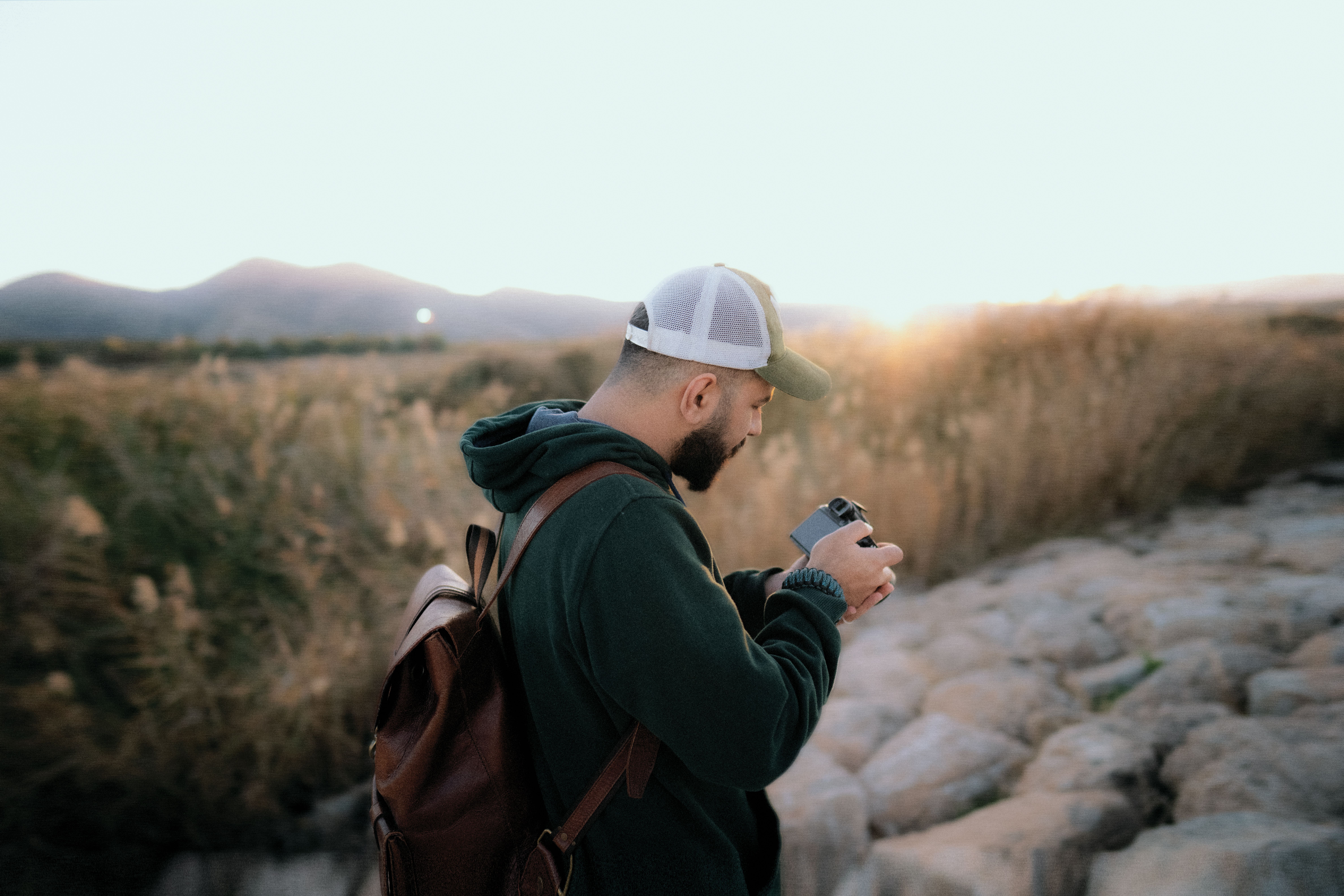 A bearded man stands on rocky terrain, capturing the serene mountain sunset with his camera.