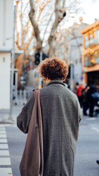 Fashionable woman with curly hair wearing a coat, standing on a bustling city street with a tote bag.