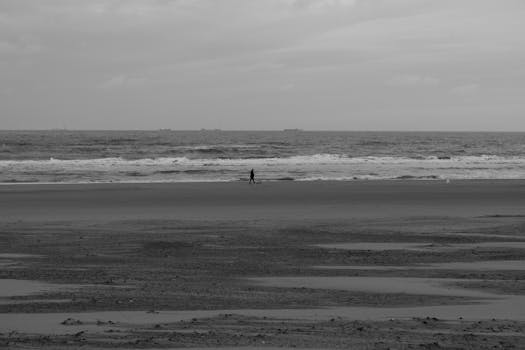 Black and white photo of a solitary figure walking by the ocean under a cloudy sky.