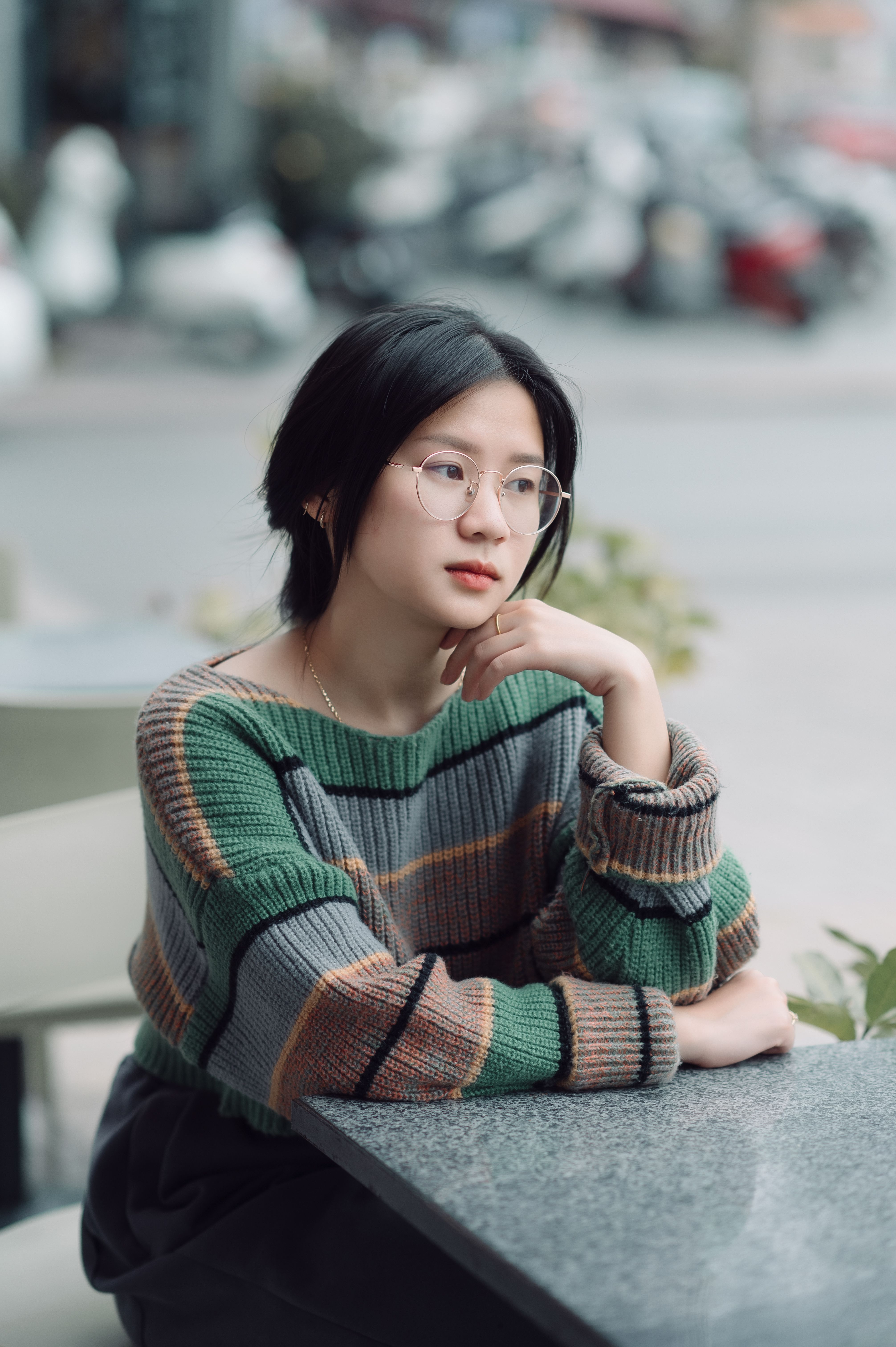 A pensive woman in eyeglasses and a colorful sweater sits at a cafe table, looking thoughtful.
