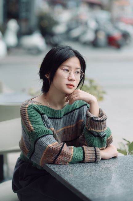 A pensive woman in eyeglasses and a colorful sweater sits at a cafe table, looking thoughtful.